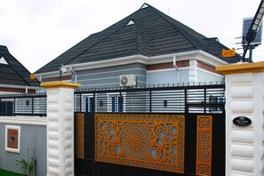 A gated entrance with intricate orange and black decorative panels, a white building with a grey roof, and a sign that reads "The Peak Residence."