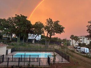 A swimming pool surrounded by a black fence, trees, and buildings in the background. - Spacieux appartement dans résidence avec piscine (Bormes-les-Mimosas)