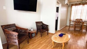 A living room with a wooden floor, a sofa, two chairs, a coffee table, and a flat-screen TV mounted on the wall. - Stylish 1-bed Opposite Queens Park Savannah (Port of Spain)
