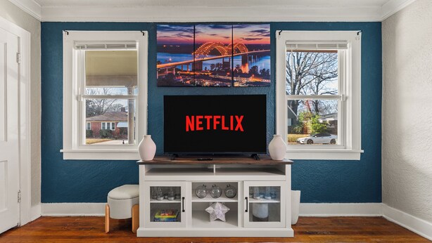 Una sala de estar con un televisor montado en un mueble blanco, dos ventanas con vistas a un puente y casas, y una pared azul.