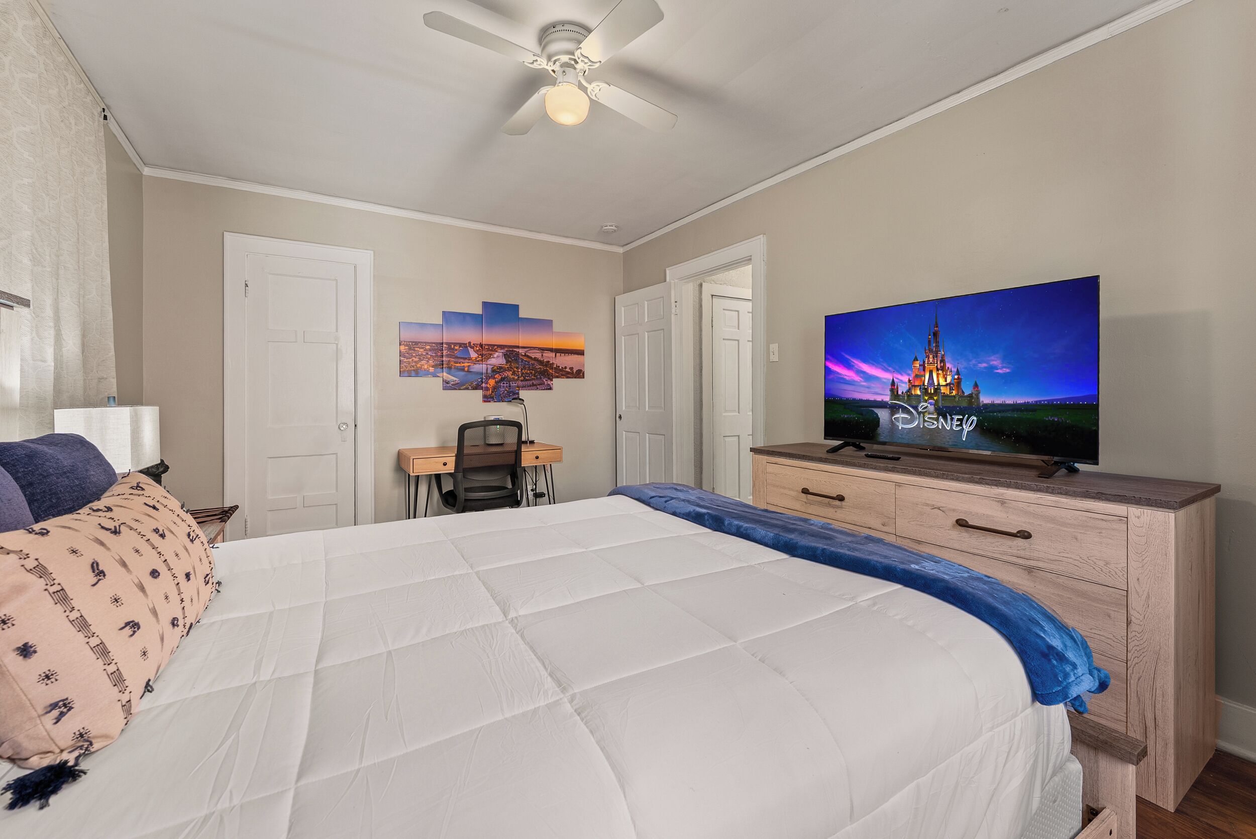 A hotel room with a bed, a desk, a chair, a dresser, and a TV displaying a Disney castle.