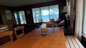 Taula de dinar de fusta amb cadires, una cistella de vimet i vista al mar a través de grans finestres.