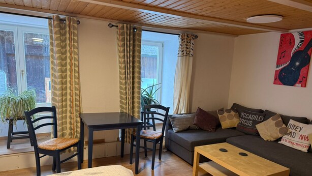 A cozy living room with a sofa, a wooden coffee table, a dining table with chairs, and a bed with a patterned duvet.