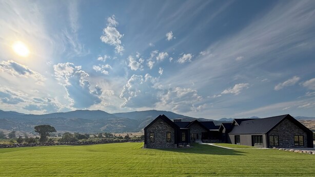 Une maison en pierre avec une grande pelouse, située dans un cadre magnifique avec des montagnes en arrière-plan et un ciel dégagé.