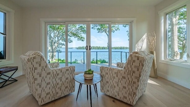 Een ruime kamer met een grote schuifpui en uitzicht op het meer. Er staan twee gestreepten fauteuils en een klein rond tafeltje met een plant.