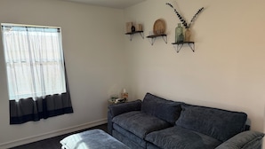 Ein Wohnzimmer mit grauem Sofa, passendem Hocker und einem Fenster mit durchscheinenden Vorhängen. An der Wand befinden sich drei Regale mit Dekoration.