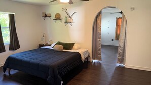 Ein Schlafzimmer mit Bett, Nachttisch, Deckenventilator, Fenster mit Vorhängen und einem zur anderen Kammer führenden Gang.