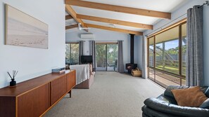 A spacious living room with a fireplace, a sofa, a TV, and a large window offering a view of the outdoors. - Lohr Beach House - Surf Side (Inverloch)