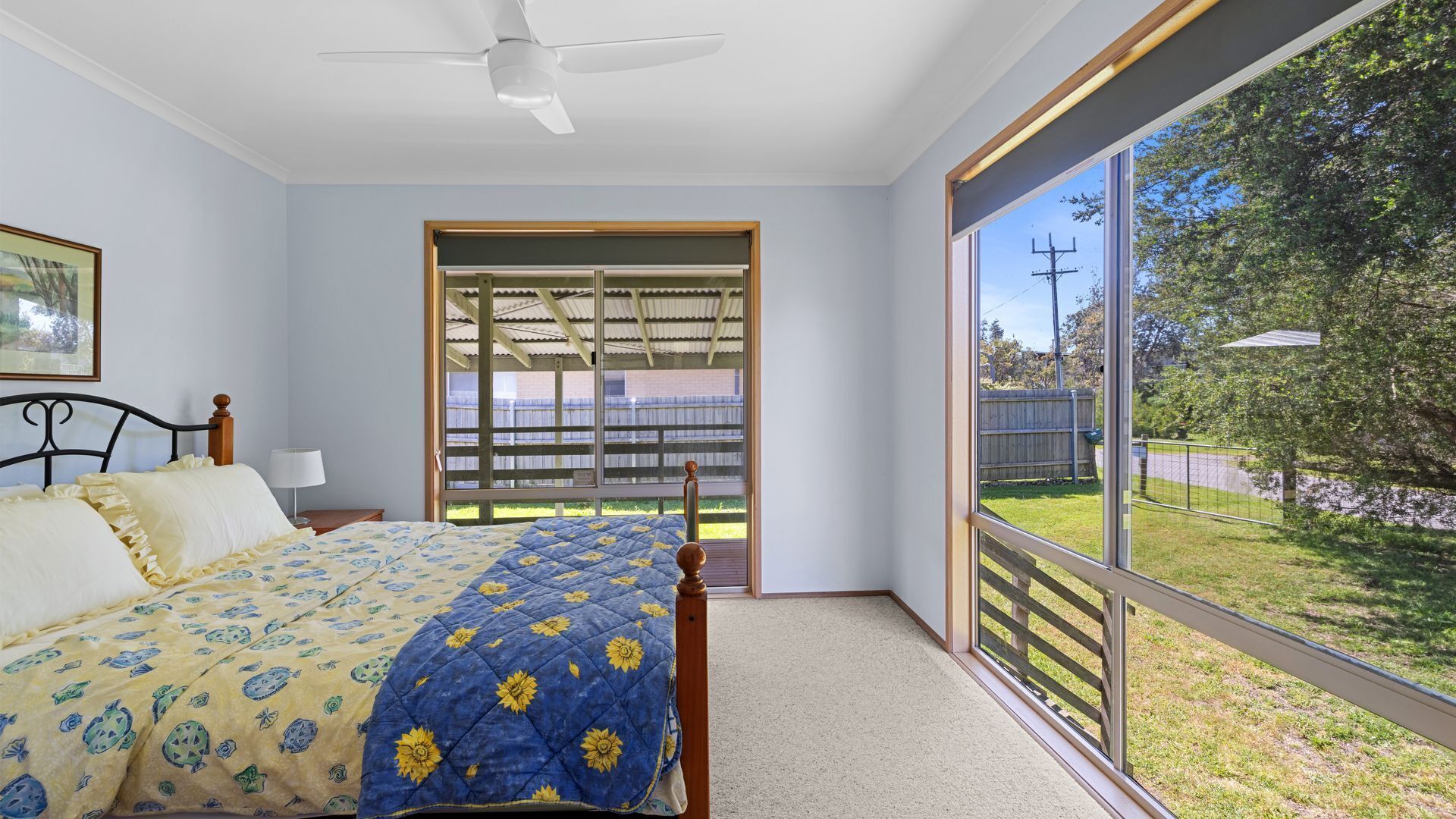 A bedroom with a bed, a ceiling fan, a large window, and a view of a backyard.