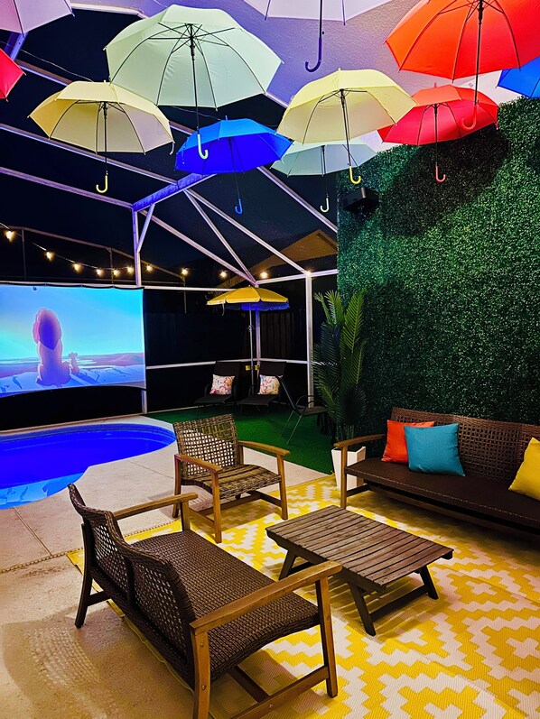 Un espace extérieur aménagé avec des meubles en osier, une piscine et un grand écran diffusant un film.