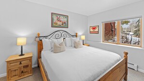 Quarto com cama de madeira, duas mesas de cabeceira com luminárias, janela com vista para árvores e duas imagens emolduradas na parede.