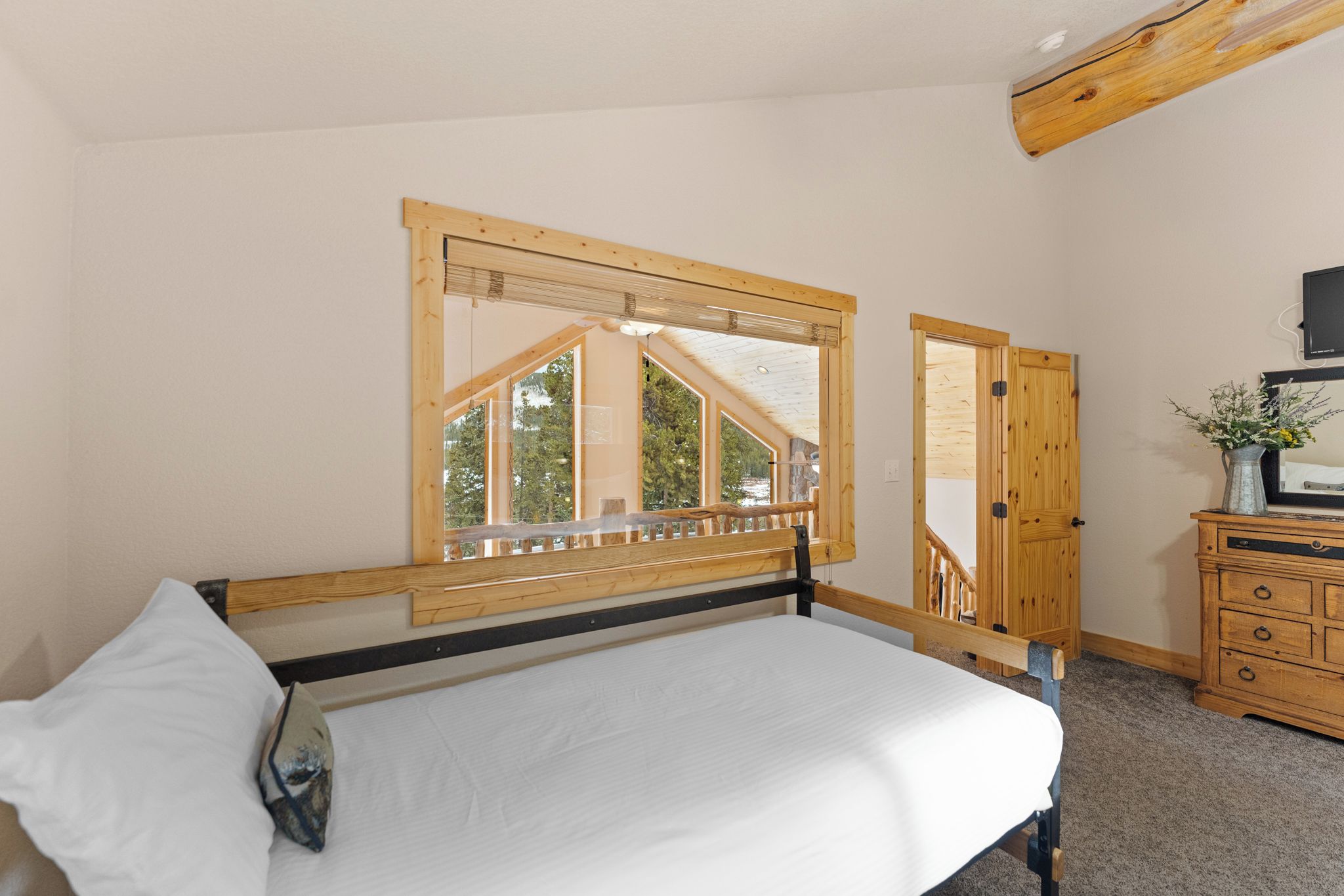 A bedroom with a bed, wooden headboard, a window with a view of trees, a wooden dresser, and a television on a stand.