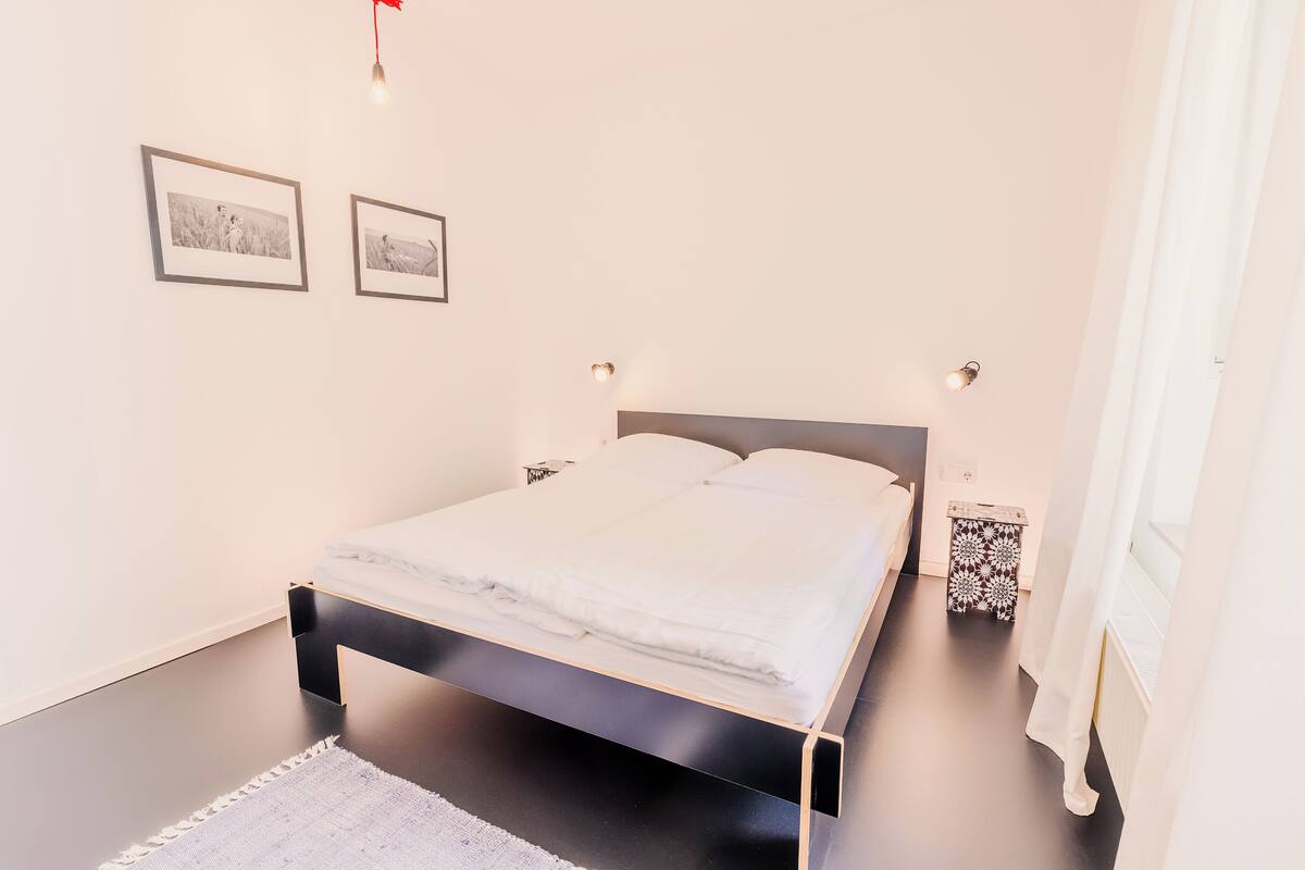 Un lit bien fait, avec du linge de lit blanc, dans une pièce dont le sol est foncé, les murs blancs et où l’on peut voir deux tableaux encadrés accrochés au mur.