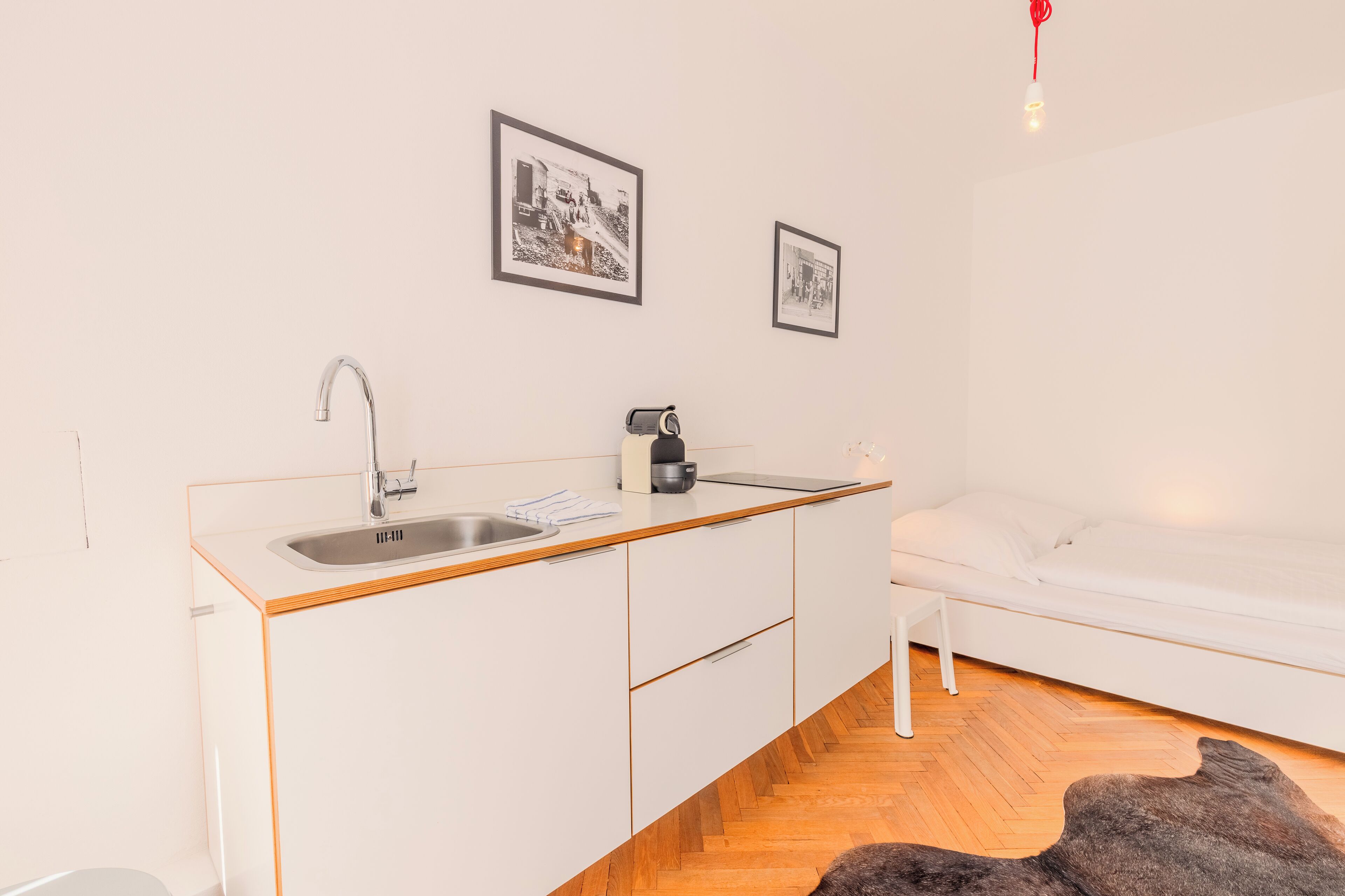 A modern, compact room with a kitchenette, a white bed, and a wooden floor.