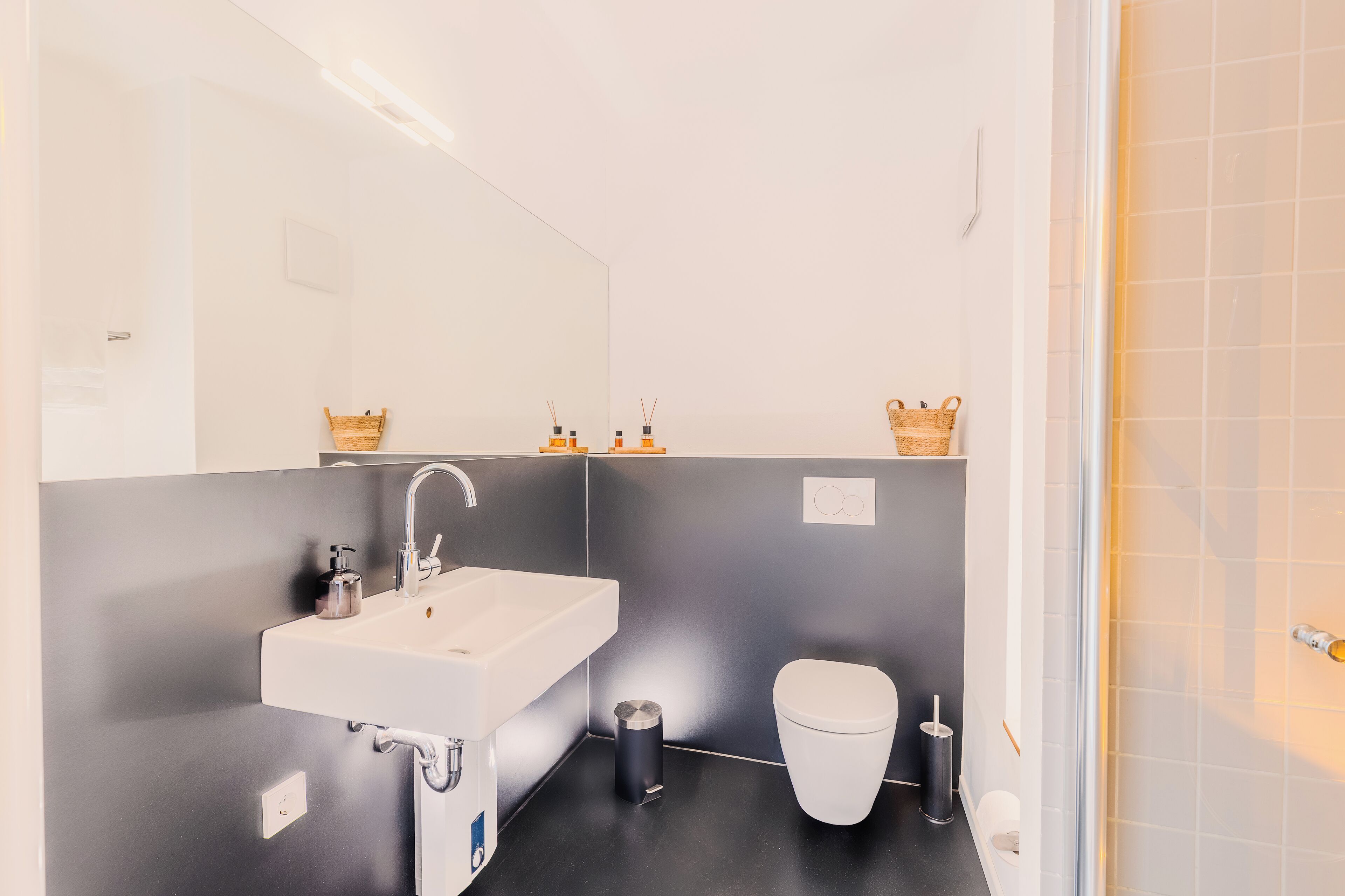 A modern bathroom with a white sink, a toilet, and a mirror. There are baskets on the wall and a trash can on the floor.