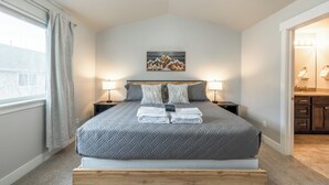 Une chambre à coucher bien rangée, avec un grand lit, deux tables de chevet avec des lampes, un tableau au mur et une vue sur la salle de bain par une porte ouverte.
