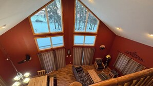 A cozy loft-style room with a sloped ceiling, large windows offering a view of a snowy landscape, a wooden staircase, and a rustic wooden bench.