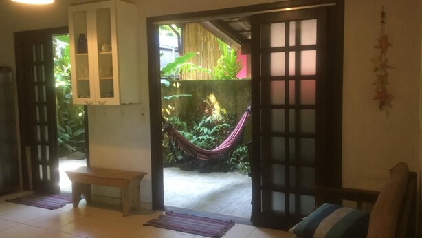 Une porte ouverte donne sur un jardin où se trouvent un hamac, un banc en bois et une armoire aux portes vitrées.