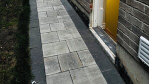 Un sendero estrecho que conduce a un edificio con una puerta amarilla, al lado de un muro de ladrillo y una zona verde.