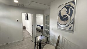 Une chambre d’hôtel moderne avec une table à manger en verre, une seule chaise, un grand tableau abstrait au mur et un lit visible dans la pièce adjacente.