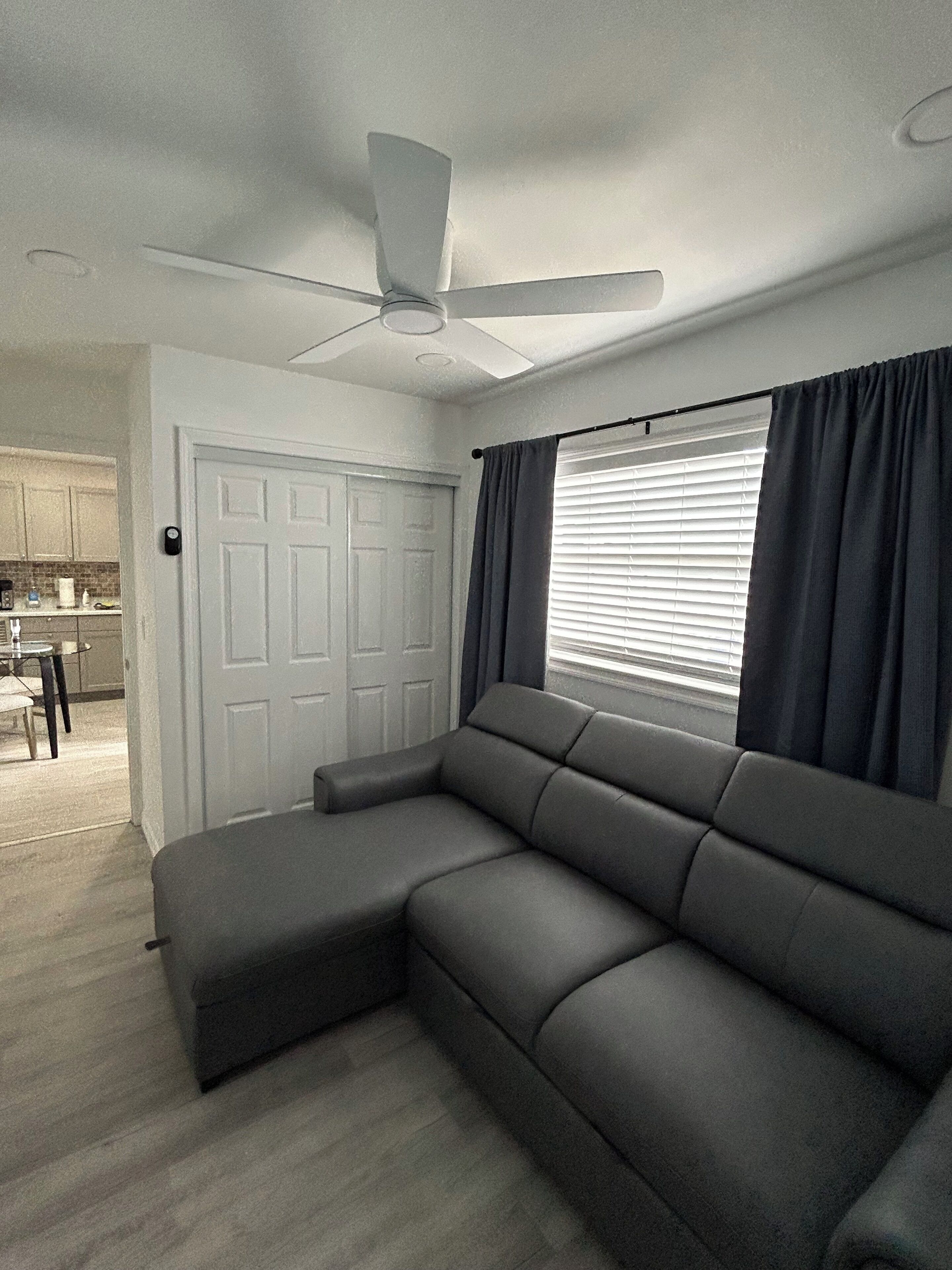 Ein modernes Wohnzimmer mit Ecksofa, Deckenventilator, einer Tür und einem Fenster mit Jalousien.