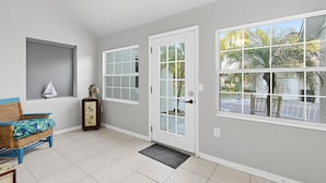 Una habitación con una puerta blanca, una ventana que da a palmeras, una silla de mimbre y una pequeña mesa con un adorno.