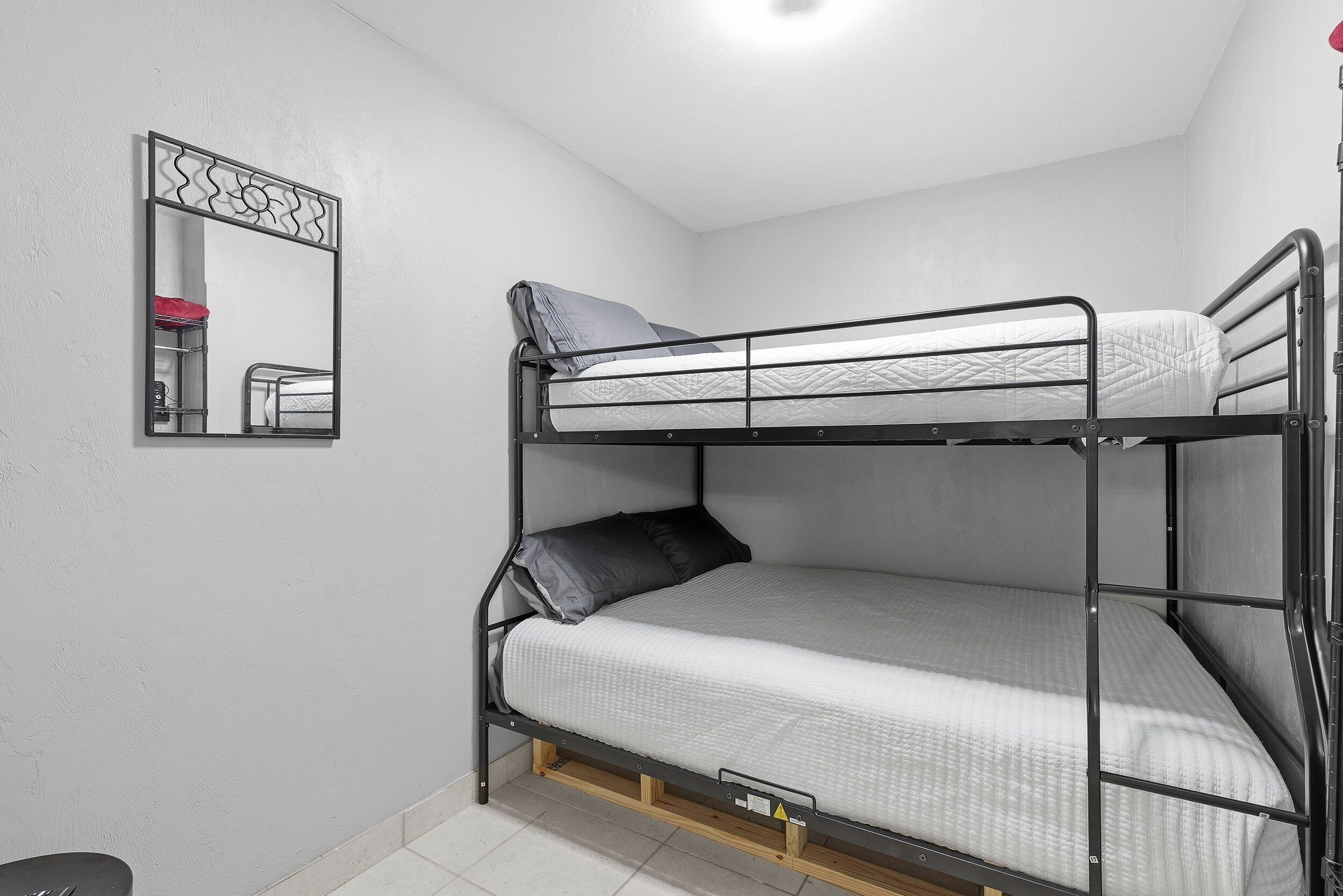 Een stapelbed met metalen frame en witte beddengoed, een spiegel met decoratieve bovenkant, en een rood voorwerp op de vloer.
