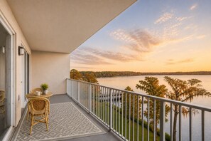 En balkon med udsigt over et søområde og træer, et bord med stole og en potteplante.