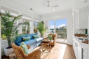 A bright, open-plan living area with a blue sofa, wicker chairs, and large windows offering a view of the outdoors. - “Roseate Tide” 9844 Haven Loop, 3 BR Kiawah River, Stunning River Views, Sleeps 6 (Johns Island)