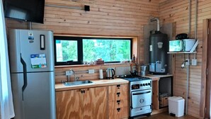 A compact kitchen with wooden cabinets, a stainless steel refrigerator, a stove, and a window overlooking greenery. - Cabaña Aida Epuyén - comfortable and bright cabin in a natural setting (Epuyén)