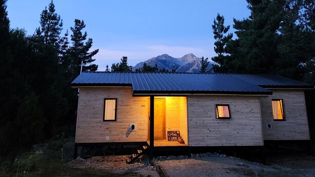 A wooden cabin with a metal roof, two windows, and a door. It is surrounded by trees and has a mountain in the background. - Cabaña Aida Epuyén - comfortable and bright cabin in a natural setting (Epuyén)