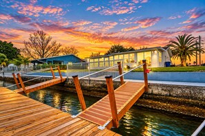 A wooden dock leading to a building with a glass facade, a palm tree, and a sunset sky. - Bay Breeze - Waterfront Retreat with Large Dock (Hernando Beach)