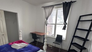 Et soveværelse med seng, skrivebord, stol, et vindue med gardiner og en sort reol.