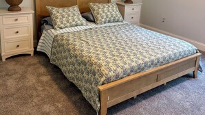 Un dormitorio con una cama de madera, dos mesitas de noche, piso alfombrado y dos cuadros en la pared.