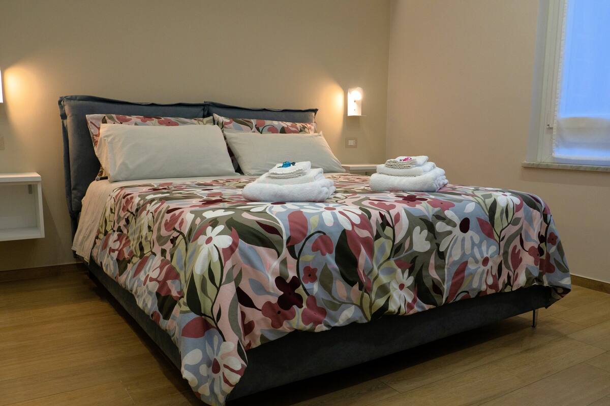 A neatly made bed with floral bedding, a bedside table, and a wall-mounted light fixture.