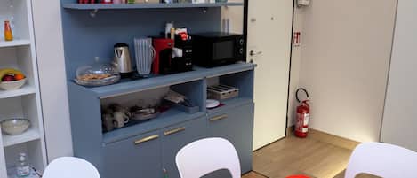 Una kitchenette compacta con microondas, cafetera y varios contenedores de almacenamiento. Hay sillas blancas y una mesita con mantel rojo. Un extintor está montado en la pared.