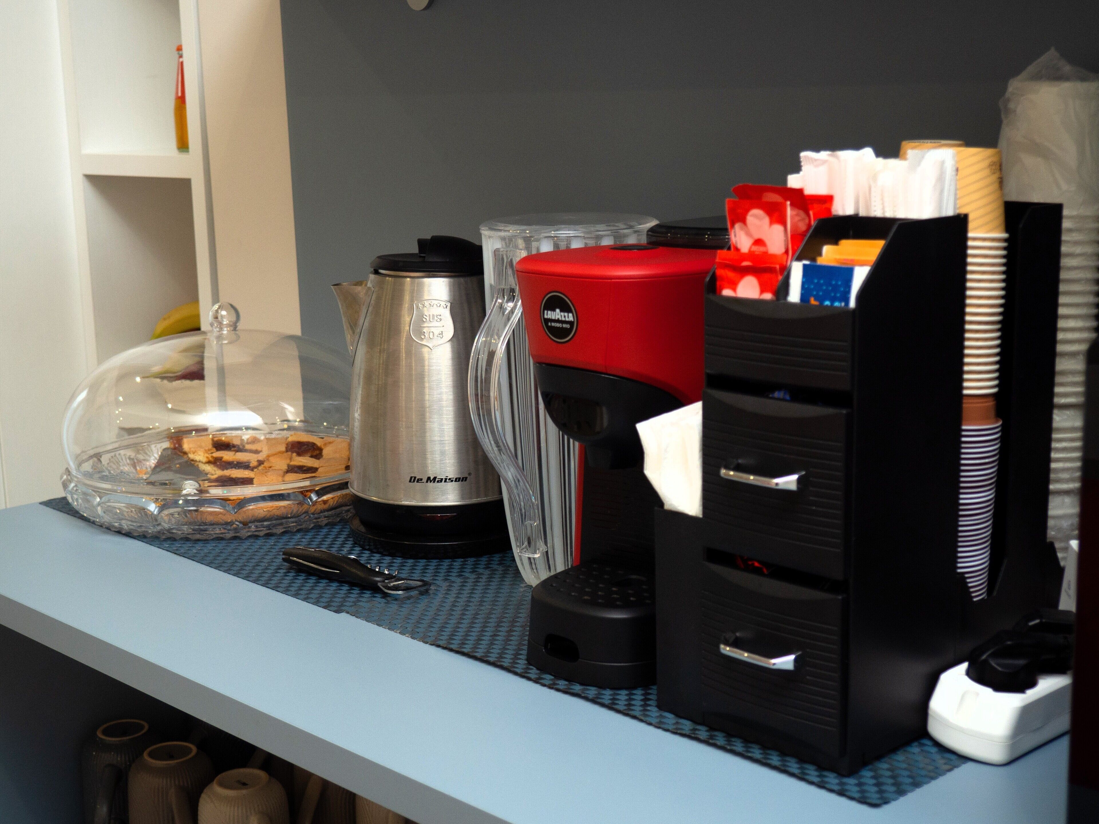 A kitchen counter with a coffee maker, a kettle, a glass container with food, and a tissue dispenser.