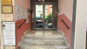 Una escalera conduce a la entrada de un edificio con varias placas en la pared, un cartel con el número '55' y vistas de coches estacionados afuera.