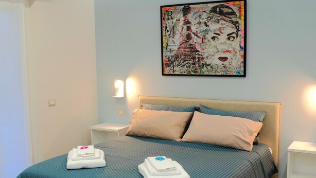 Una cama bien hecha con un edredón verde azulado y dos almohadas, un cuadro abstracto de gran tamaño en la pared, y una lámpara de noche a cada lado.
