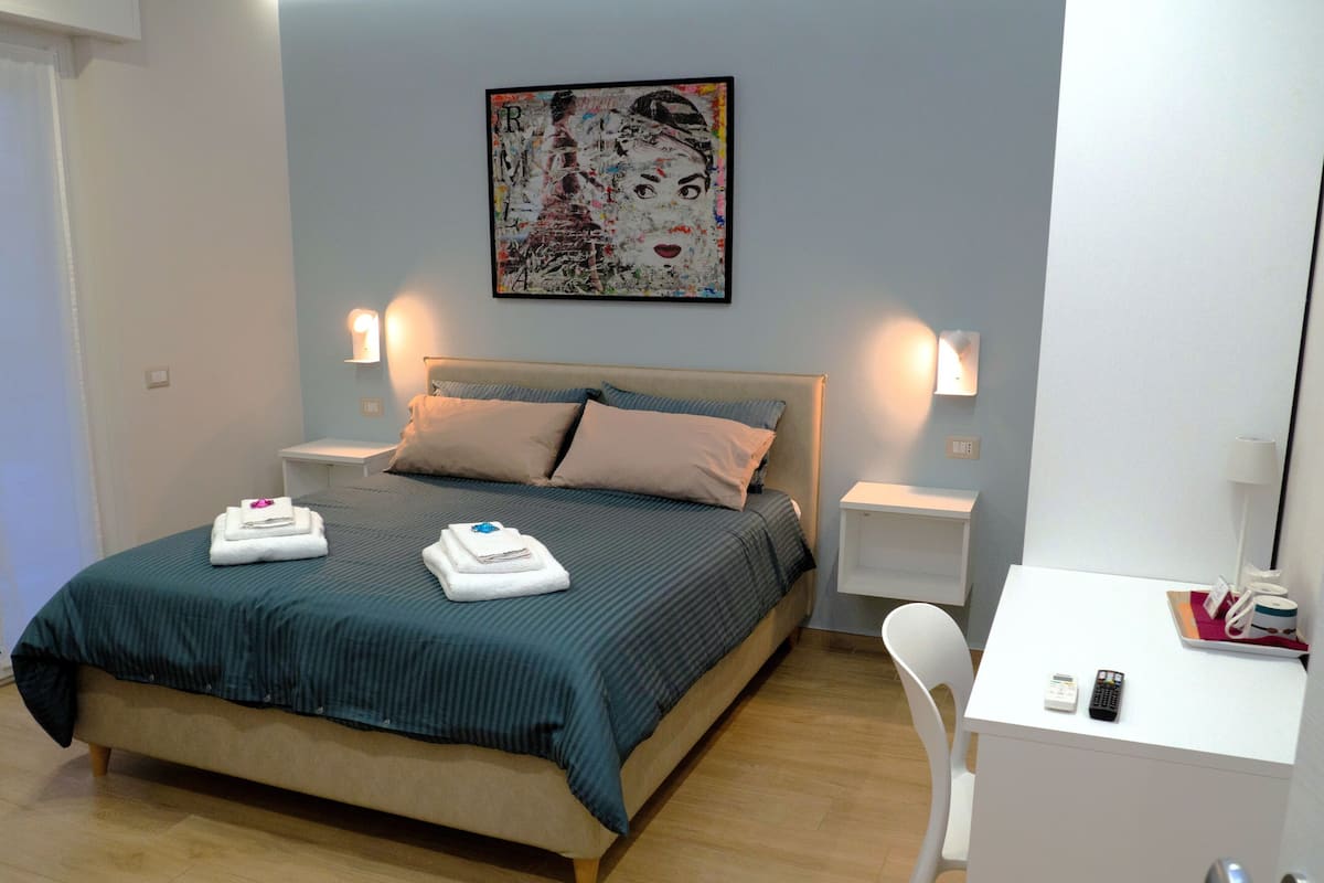 A neatly made bed with a green bedspread and two folded towels, a bedside table with a lamp, a small white desk with a phone and remote, and a painting on the wall.