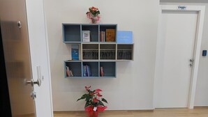 Una habitación con piso de madera, una estantería con varios objetos, una puerta que dice "Viaggio" y una pequeña planta en maceta.