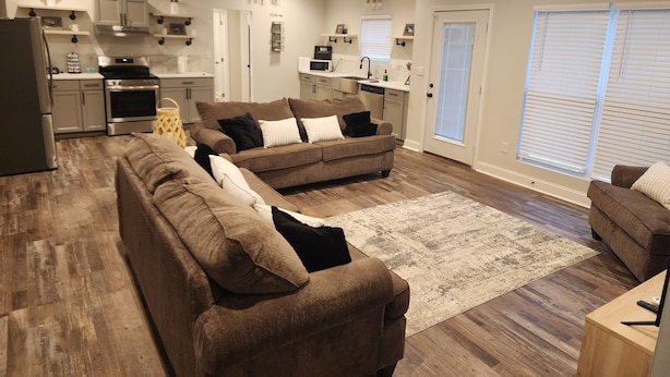 A spacious living room with two brown sofas, a wooden floor, and a kitchen area with white cabinets and a microwave.
