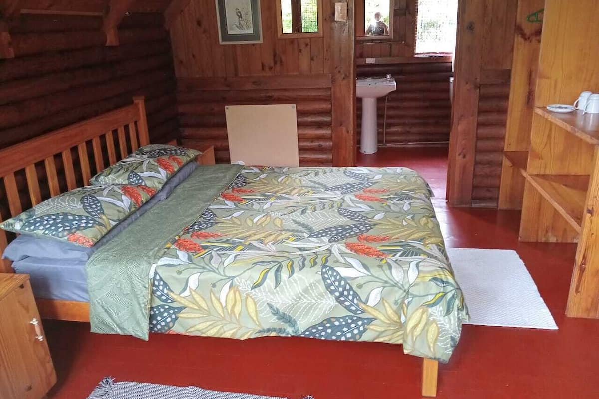 Acogedora habitación tipo cabaña de madera con una cama, un banco de madera, una mesita y un baño con lavamanos y espejo.