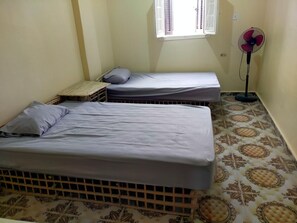 Een kleine kamer met twee eenpersoonsbedden, een verticale ventilator en een raam met luiken.