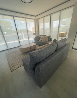 A spacious living room with a grey sofa, a coffee table, and a lamp. Large windows provide a view of the outside.