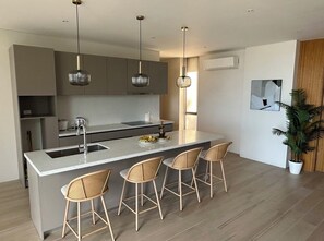 A modern kitchen with a central island, bar stools, pendant lights, and a potted plant.
