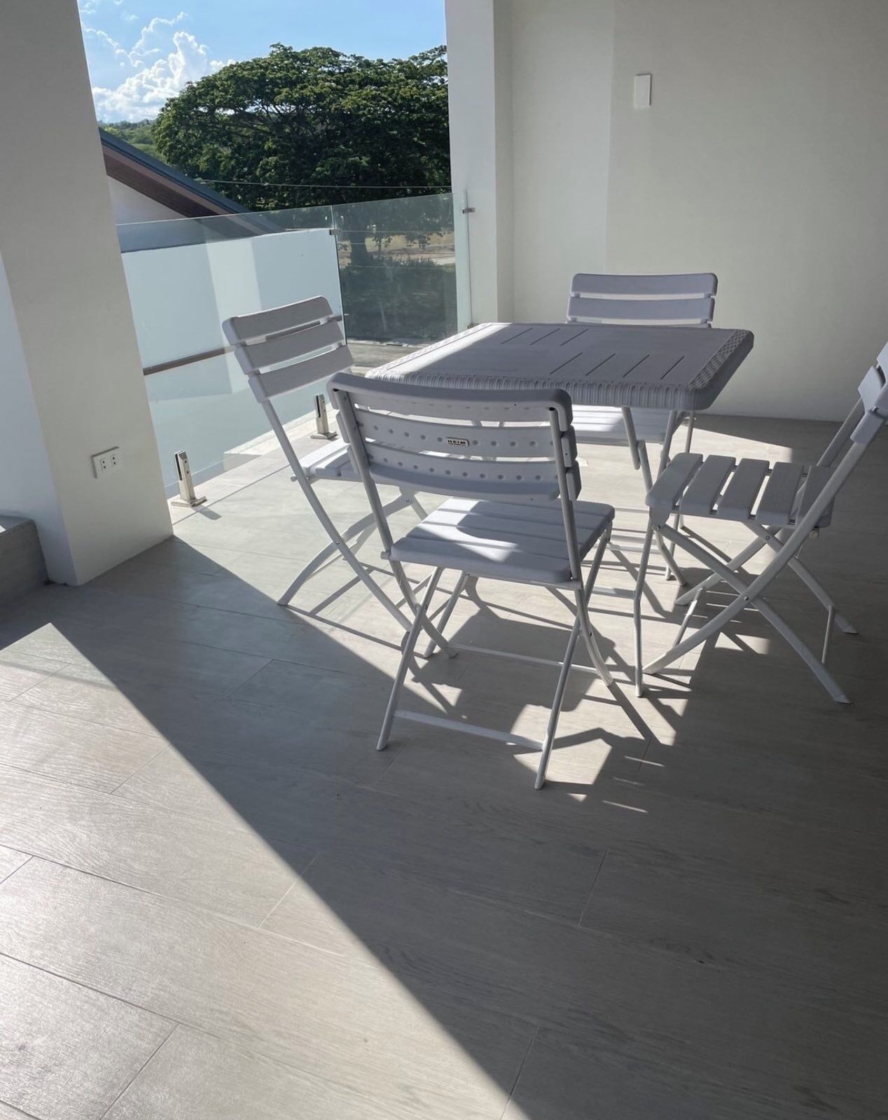 A sunlit outdoor patio with a metal table and chairs, a glass balustrade, and a view of greenery.