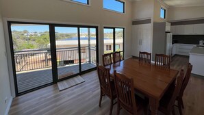 A spacious dining area with a large wooden table and chairs, a kitchen with white cabinets, and a balcony with a view of the landscape.
