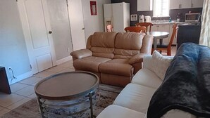 A living room with a brown leather sofa, a white sofa, a round coffee table, and a dining area with a table and chairs. - GpL 3115 B 3 Bed 1 Bath (Fort Myers)