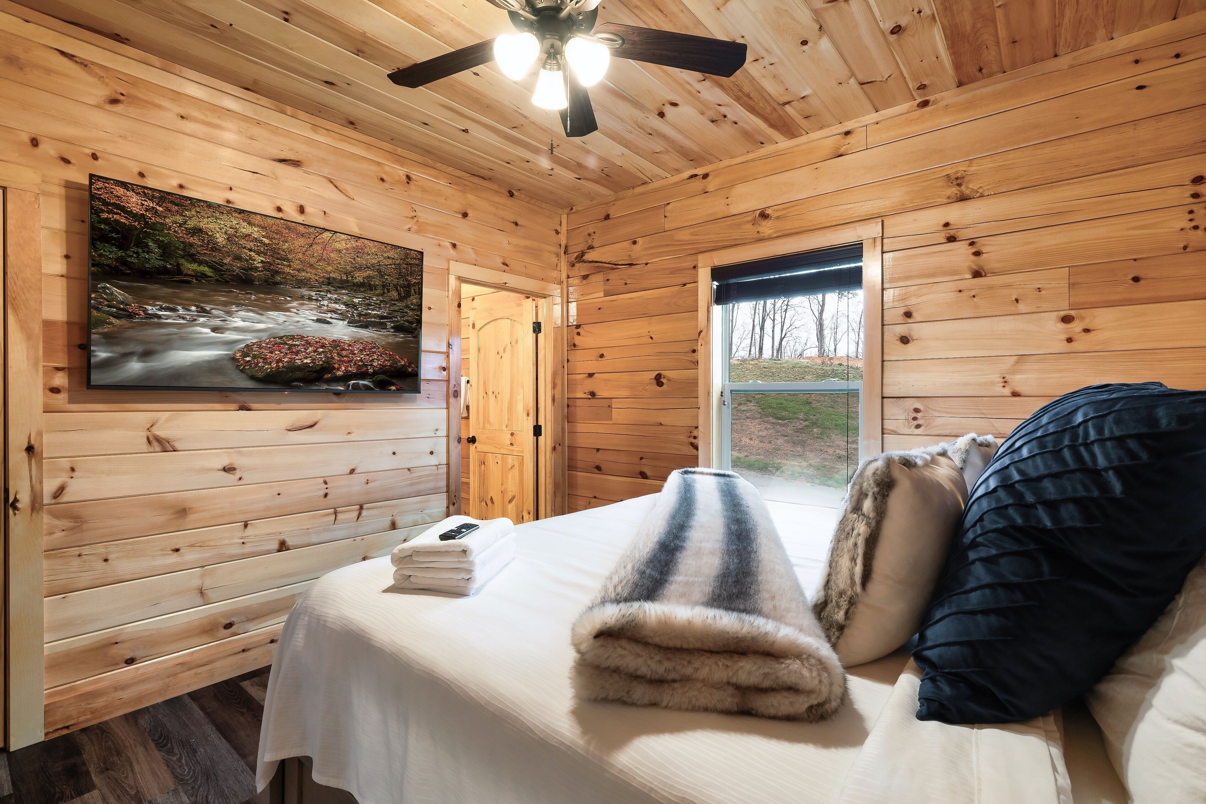 A wooden cabin room with a bed, a TV on the wall, and a window with a view of trees.